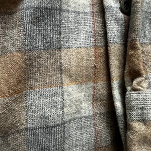 May include: Close-up of a gray, brown, and blue plaid shirt. The fabric has a textured appearance with a button and a small red stripe. The shirt is made of a woven material.