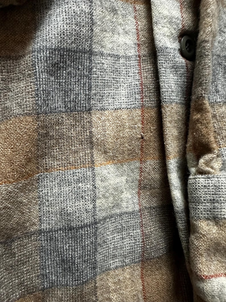 May include: Close-up of a gray, brown, and blue plaid shirt. The fabric has a textured appearance with a button and a small red stripe. The shirt is made of a woven material.