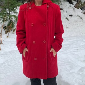 May include: A red double-breasted wool coat with a collar that can be worn up or down. The coat has four buttons down the front and two pockets on the sides.
