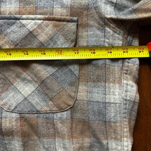 May include: Close-up of a brown, gray, and blue plaid shirt with a pocket. A yellow tape measure shows the pocket is approximately 16 inches wide. The shirt has a classic plaid pattern.