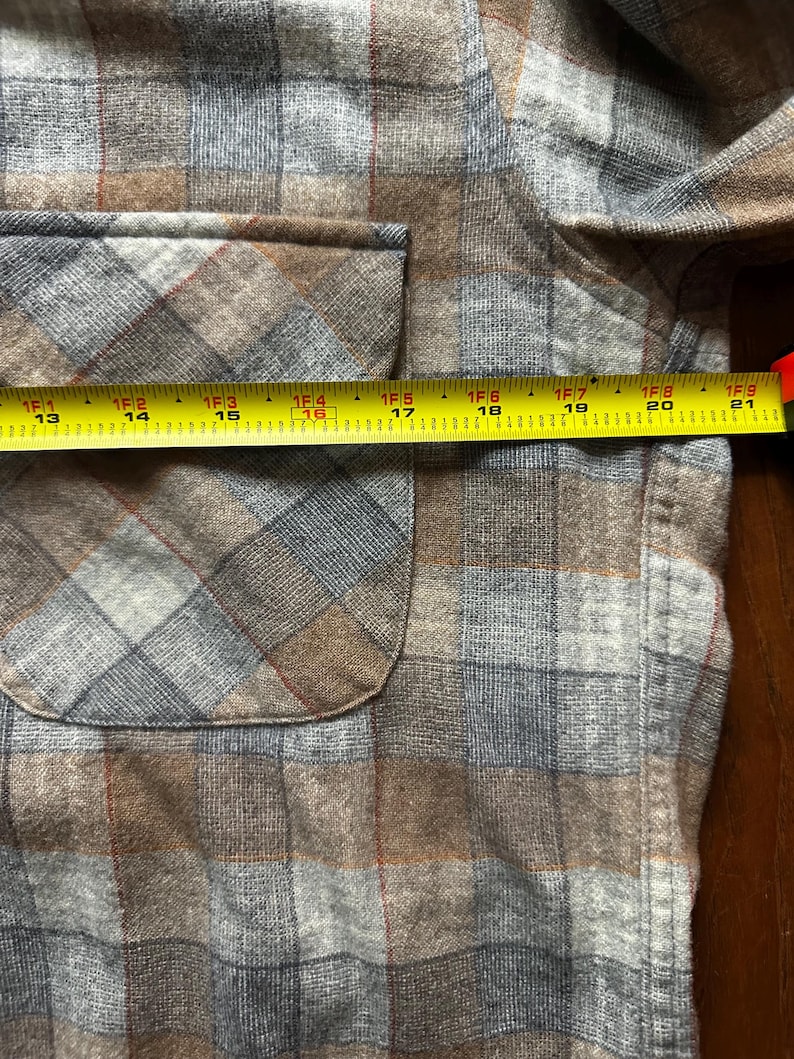 May include: Close-up of a brown, gray, and blue plaid shirt with a pocket. A yellow tape measure shows the pocket is approximately 16 inches wide. The shirt has a classic plaid pattern.