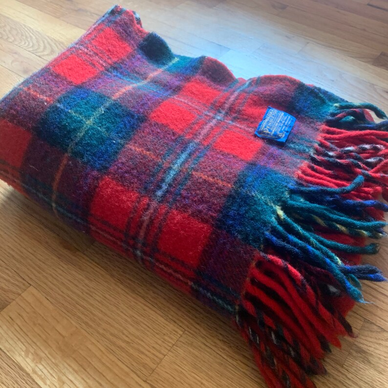 Op de afbeelding: Een rood, blauw en groen geruit wollen deken met franjes. Het deken heeft een blauw label met witte tekst dat "Pendleton" zegt.