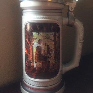 May include: A white ceramic beer stein with a silver lid and a handle. The stein features a colorful image of a blacksmith working at an anvil with a dog by his side.