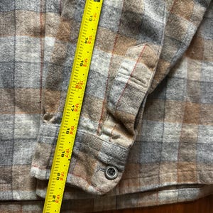 May include: A close-up of a button-down flannel shirt in a plaid pattern of brown, gray, and red. A yellow measuring tape is placed along the shirt's sleeve, showing measurements up to 23 inches.