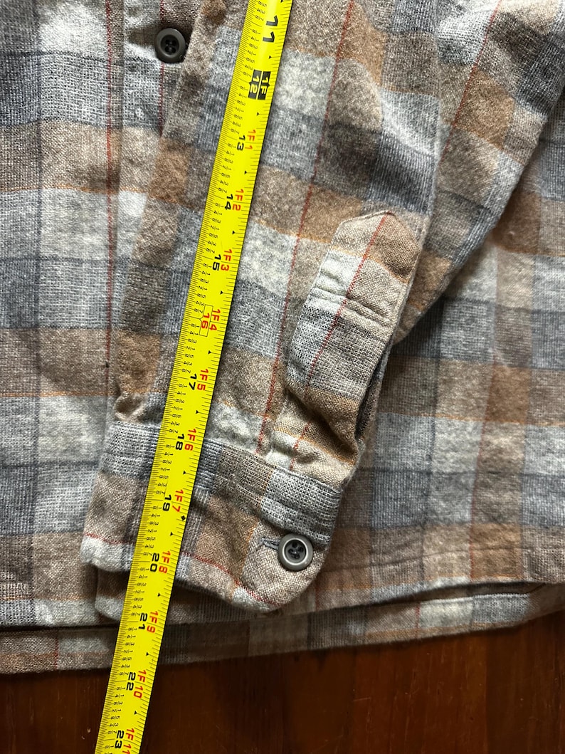 May include: A close-up of a button-down flannel shirt in a plaid pattern of brown, gray, and red. A yellow measuring tape is placed along the shirt's sleeve, showing measurements up to 23 inches.