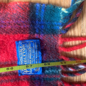 Op de afbeelding: Een rood, blauw en groen geruit Pendleton wollen deken met franjes. Het deken heeft een blauw label met de tekst "ANTED TO BE A PENDLETON MARK REG US PAT OFF PENTON WOOLEN MILLS LANG 322602 JURE VIRGIN WOOL AS A 57"
