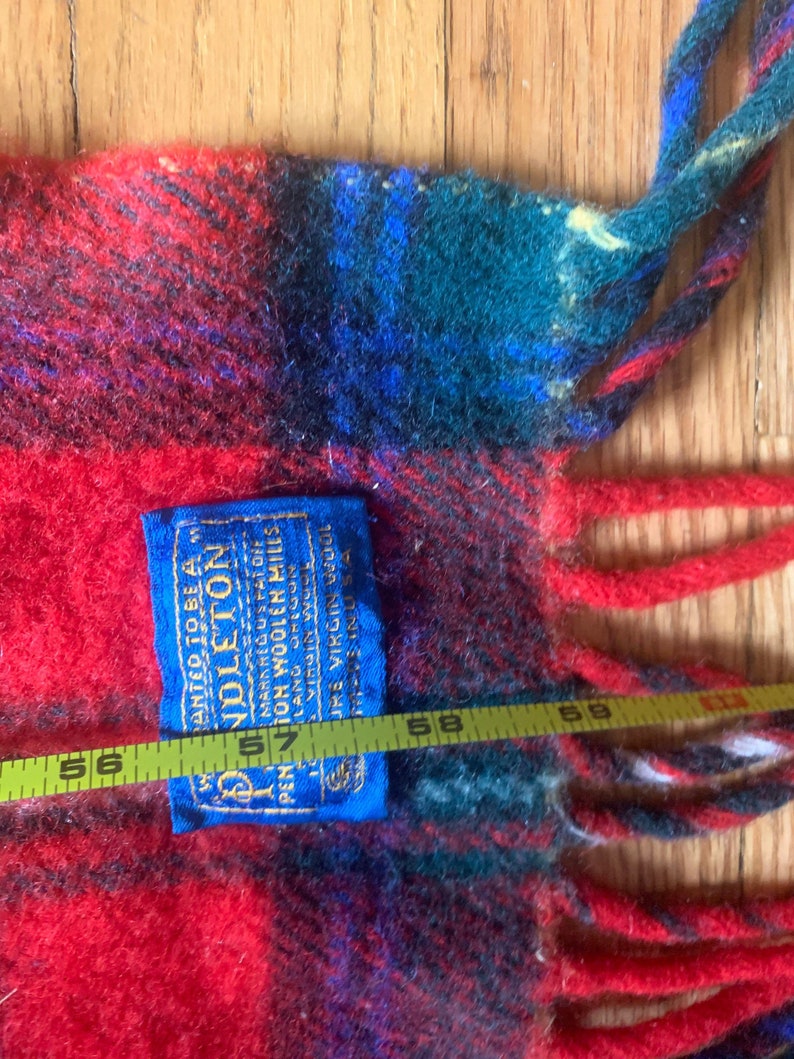 Op de afbeelding: Een rood, blauw en groen geruit Pendleton wollen deken met franjes. Het deken heeft een blauw label met de tekst "ANTED TO BE A PENDLETON MARK REG US PAT OFF PENTON WOOLEN MILLS LANG 322602 JURE VIRGIN WOOL AS A 57"