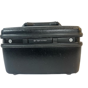 Puede incluir: Un maletín negro Samsonite con una superficie texturizada. El maletín tiene un asa en la parte superior y dos cerraduras de combinación en la parte delantera. El logotipo de Samsonite es visible.