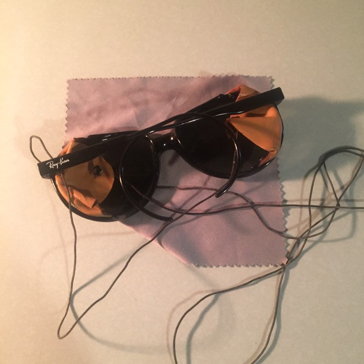 Może przedstawiać: Czarne okulary przeciwsłoneczne Ray-Ban z pękniętą soczewką i fioletowym ściereczką do czyszczenia. Okulary przeciwsłoneczne leżą na białym tle z czarnym sznurkiem.