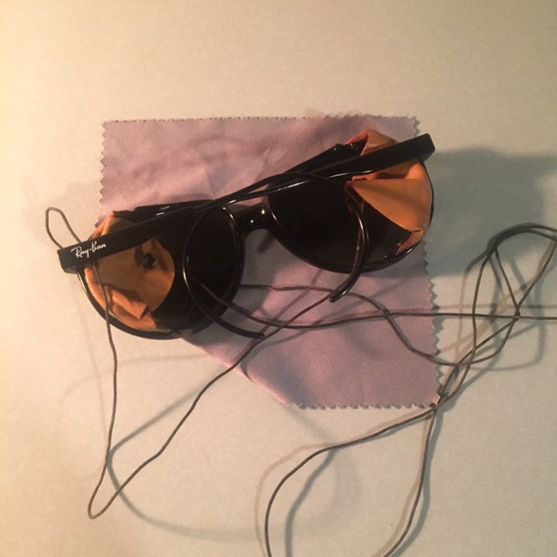 Może przedstawiać: Czarne okulary przeciwsłoneczne Ray-Ban z pękniętą soczewką i fioletowym ściereczką do czyszczenia. Okulary przeciwsłoneczne leżą na białym tle z czarnym sznurkiem.