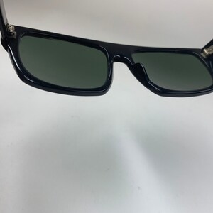 Peut inclure: Lunettes de soleil noires avec une monture rectangulaire et des verres vert fonc&eacute;. Les lunettes sont ouvertes et les charni&egrave;res sont visibles.