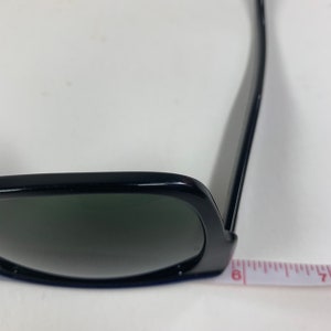 Peut inclure: Lunettes de soleil noires avec des verres vert fonc&eacute; et une monture noire. Les lunettes de soleil reposent sur une surface blanche avec un ruban &agrave; mesurer.