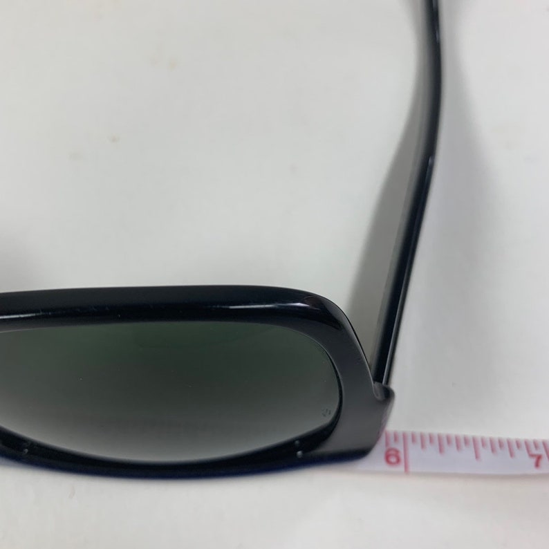 Peut inclure: Lunettes de soleil noires avec des verres vert fonc&eacute; et une monture noire. Les lunettes de soleil reposent sur une surface blanche avec un ruban &agrave; mesurer.