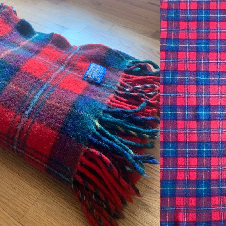 Op de afbeelding: Een rode, blauwe en groene geruite deken met franjes. De deken heeft een blauw label met de tekst "Pendleton."