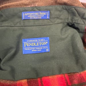 以下が含まれることがあります： 緑と茶色の格子柄のペンドルトンシャツ。青いラベルには「Adventure Shirt Assembled in Mexico of USA Virgin Wool」と「Warranted to be a Pendleton Pendleton Woolen Mills Since 1863」と書いてあります。
