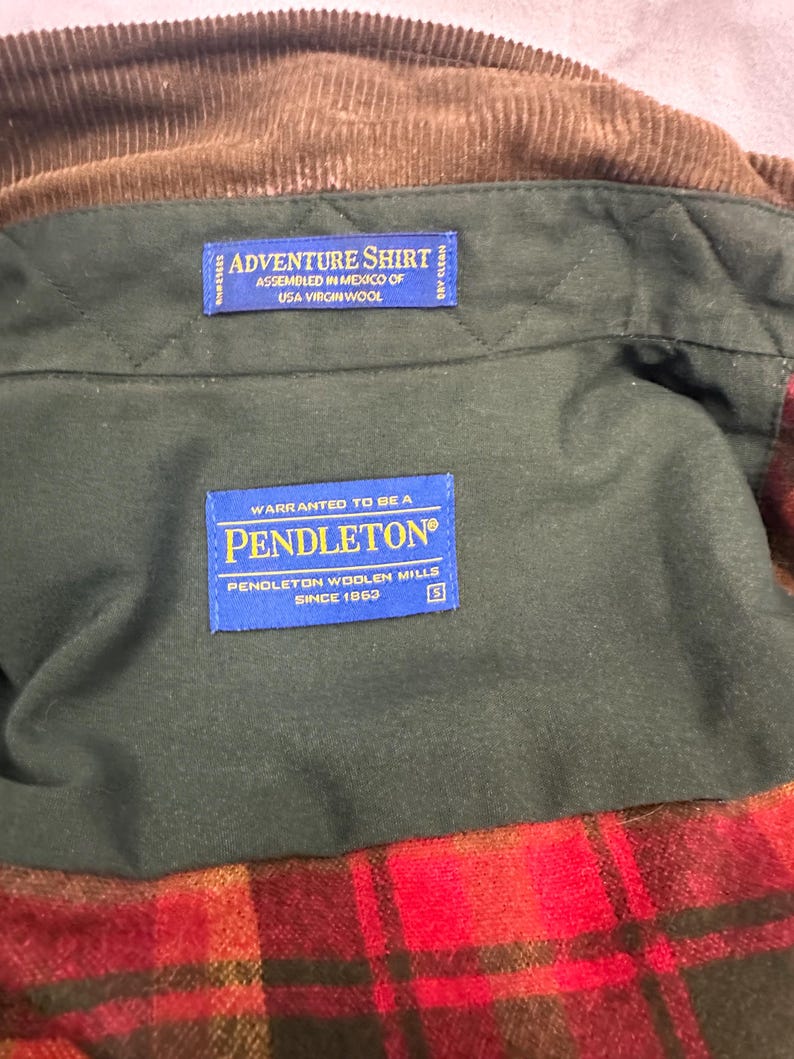 以下が含まれることがあります： 緑と茶色の格子柄のペンドルトンシャツ。青いラベルには「Adventure Shirt Assembled in Mexico of USA Virgin Wool」と「Warranted to be a Pendleton Pendleton Woolen Mills Since 1863」と書いてあります。