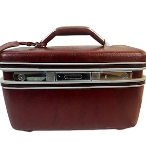 Puede incluir: Una maleta de viaje burdeos vintage con asa y adornos plateados. La maleta tiene un asa de cuero y un cierre con cerradura. La maleta es rectangular con bordes redondeados y un ribete blanco.