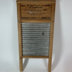 Op de afbeelding: Een vintage houten wasbord met een gegolfd metalen oppervlak. Het wasbord heeft een houten frame en twee poten. Het wasbord heeft een label met de tekst "DEAL FOR SILKS, HOSIERY AND LINGERIE OR HANDKERCHIEFS JUST THE RIGHT SIZE TO FIT A BUCKET PAIL OR LAVATORY PACKS EASILY INTO SUIT CASE OR TRAVELING BAG COLUMBUS WASHBOARD CO., COLUMBUS, OHIO".