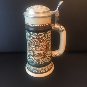Puede incluir: Una jarra de cerveza de cerámica con un diseño verde y marrón que representa una escena de caza con un perro y un cazador. La jarra tiene una tapa de metal con asa y el texto "Point...English Setter" en la parte inferior.