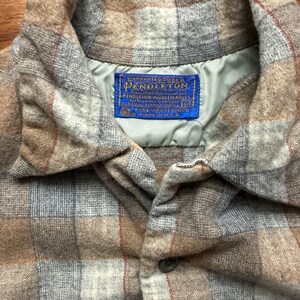 May include: Close-up of a plaid button-down shirt in shades of brown, gray, and red. The shirt features a collar and a button closure. A blue label with the text "Pendleton" is visible.