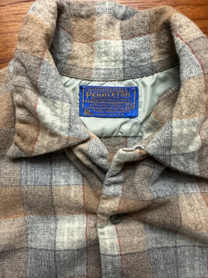May include: Close-up of a plaid button-down shirt in shades of brown, gray, and red. The shirt features a collar and a button closure. A blue label with the text "Pendleton" is visible.