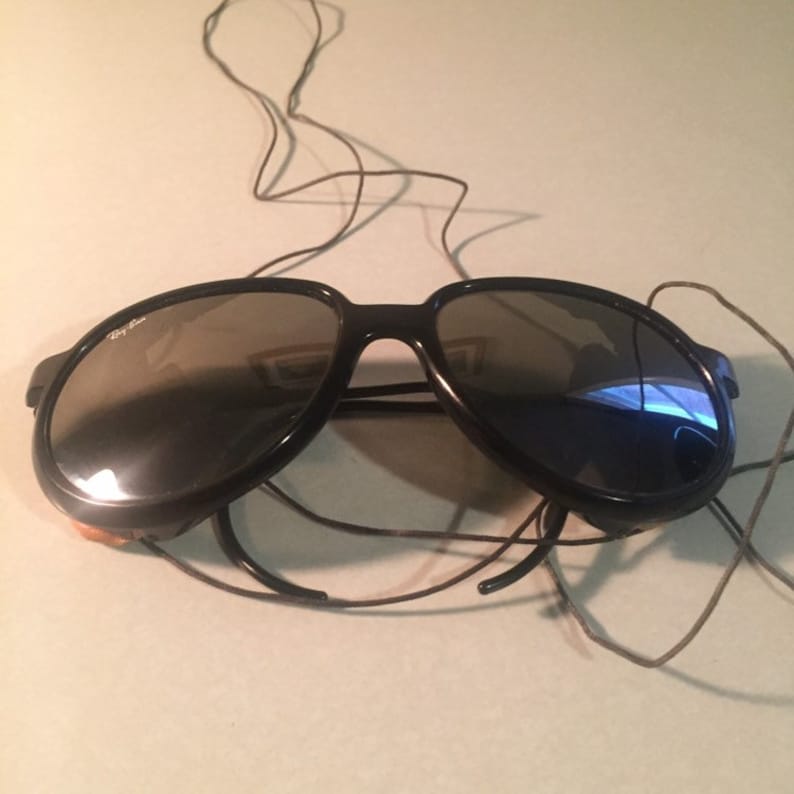 Może przedstawiać: Czarne okulary przeciwsłoneczne typu aviator z ciemnymi szkłami i czarnym sznurkiem.
