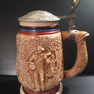 May include: A ceramic beer stein with a hinged metal lid. The stein features a relief design of a country and western music scene, with a cowboy playing a banjo. The handle is wood-toned with a decorative design.