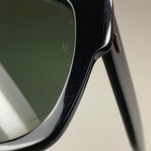 Peut inclure: Gros plan sur la lentille gauche de lunettes de soleil noires avec des verres verts. Le logo Ray-Ban est grav&eacute; sur la lentille.