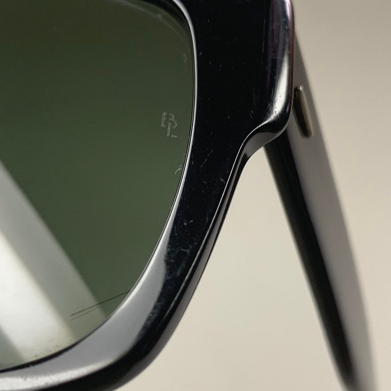 Peut inclure: Gros plan sur la lentille gauche de lunettes de soleil noires avec des verres verts. Le logo Ray-Ban est grav&eacute; sur la lentille.