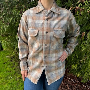 May include: A button-down plaid shirt in shades of tan, gray, and light brown. The shirt has two chest pockets and is worn with blue jeans. The shirt's fabric appears to be a soft, woven material.