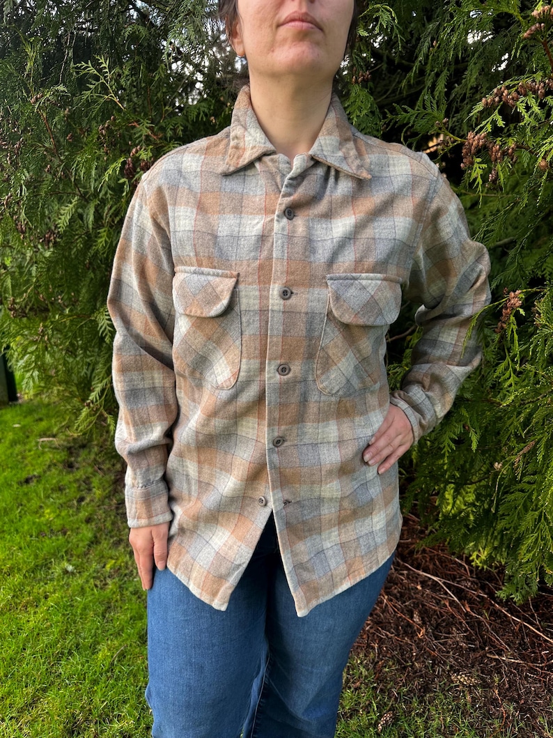 May include: A button-down plaid shirt in shades of tan, gray, and light brown. The shirt has two chest pockets and is worn with blue jeans. The shirt's fabric appears to be a soft, woven material.