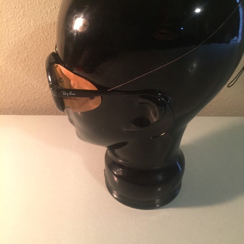 Może przedstawiać: Czarna plastikowa głowa manekina w czarnych okularach przeciwsłonecznych Ray-Ban ze złotymi szkłami. Okulary mają logo Ray-Ban z boku.