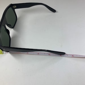 Peut inclure: Lunettes de soleil noires avec une monture &eacute;paisse et des verres fonc&eacute;s. Les lunettes sont mesur&eacute;es avec une r&egrave;gle, montrant que la longueur du cadre est d'environ 14 cm.