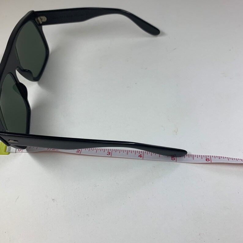 Peut inclure: Lunettes de soleil noires avec une monture &eacute;paisse et des verres fonc&eacute;s. Les lunettes sont mesur&eacute;es avec une r&egrave;gle, montrant que la longueur du cadre est d'environ 14 cm.