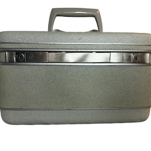 Puede incluir: Una maleta gris vintage con una superficie texturizada y un asa gris. La maleta tiene forma rectangular con detalles metálicos plateados y un mecanismo de cierre seguro. La maleta está diseñada para viajar.