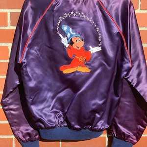 May include: A purple satin bomber jacket with a red embroidered Mickey Mouse design on the back. Mickey is dressed as a sorcerer and is surrounded by stars. The jacket has a blue ribbed collar and cuffs.