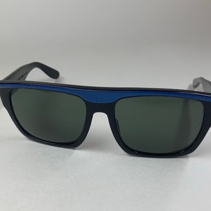 Peut inclure: Lunettes de soleil noires avec une bordure bleue autour des verres. Les lunettes ont une forme rectangulaire et des verres gris fonc&eacute;.