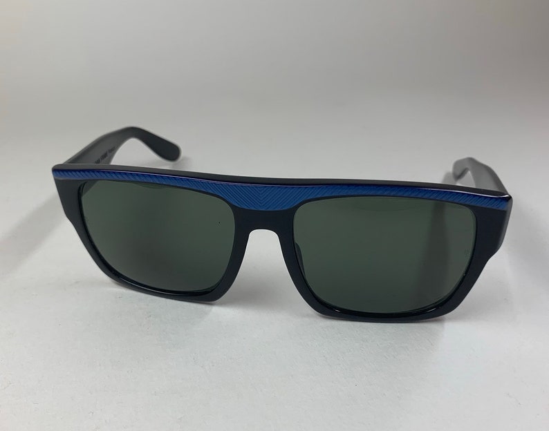 Peut inclure: Lunettes de soleil noires avec une bordure bleue autour des verres. Les lunettes ont une forme rectangulaire et des verres gris fonc&eacute;.