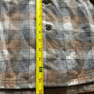May include: A close-up of a brown, gray, and tan plaid shirt with two buttons and a pocket. A yellow measuring tape is placed vertically along the shirt, showing measurements from 17 inches to 27 inches.