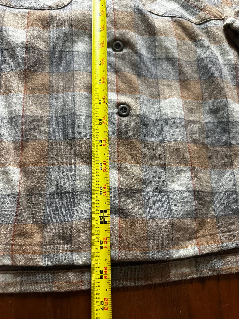 May include: A close-up of a brown, gray, and tan plaid shirt with two buttons and a pocket. A yellow measuring tape is placed vertically along the shirt, showing measurements from 17 inches to 27 inches.