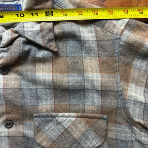 May include: A close-up of a plaid shirt in shades of gray, brown, and red. The shirt has a button-down front, a chest pocket, and a collar. The shirt is measured with a tape measure, showing a length of approximately 10 inches.