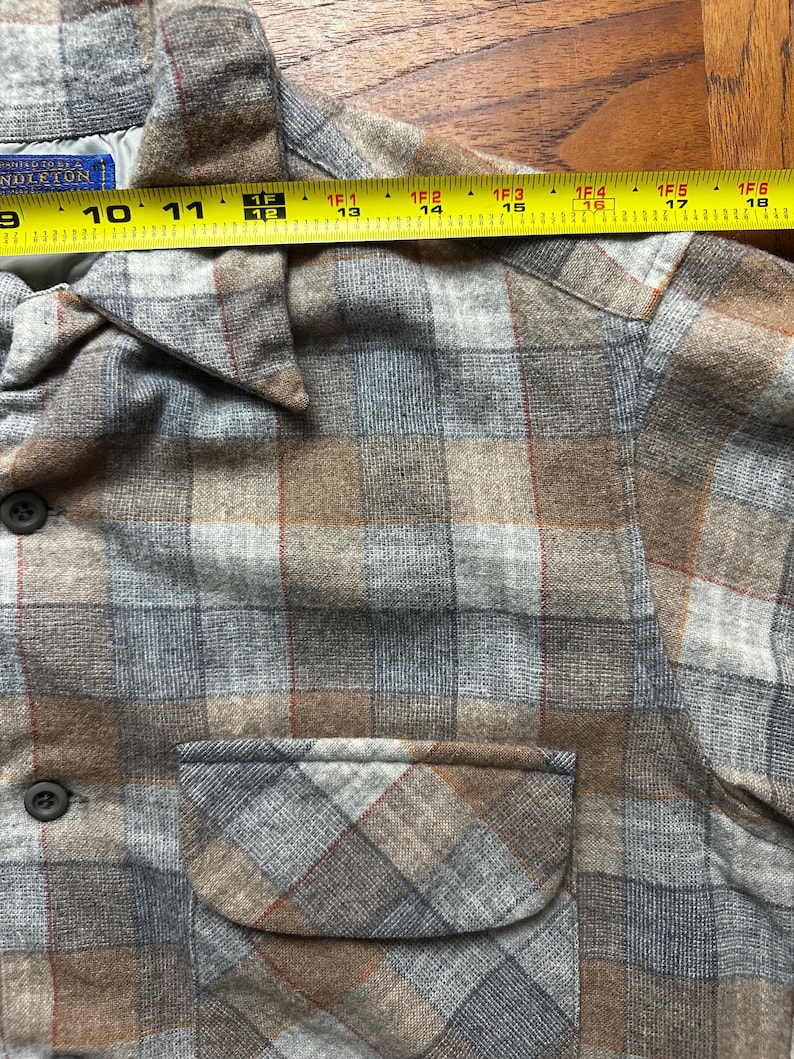 May include: A close-up of a plaid shirt in shades of gray, brown, and red. The shirt has a button-down front, a chest pocket, and a collar. The shirt is measured with a tape measure, showing a length of approximately 10 inches.