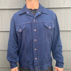 Könnte beinhalten: Eine dunkelblaue Jeansjacke mit Knopfleiste und zwei Brusttaschen. Die Jacke hat einen klassischen Stil und ist aus robustem Material gefertigt.