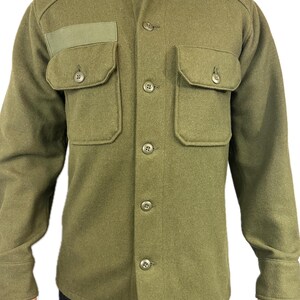 Puede incluir: Una camisa verde de manga larga con cierre de botones y dos bolsillos grandes. La camisa tiene un parche en el pecho izquierdo.