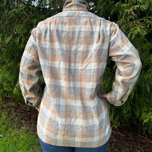 May include: A plaid button-down shirt in shades of tan, gray, and blue. The long-sleeved shirt has a collared neckline and is paired with blue jeans. The back of the shirt is visible, showcasing the plaid pattern.