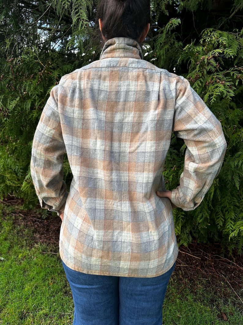May include: A plaid button-down shirt in shades of tan, gray, and blue. The long-sleeved shirt has a collared neckline and is paired with blue jeans. The back of the shirt is visible, showcasing the plaid pattern.