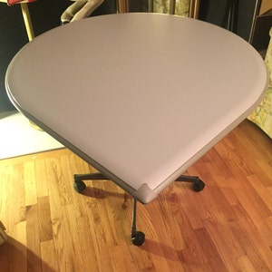 May include: A white, round table with a unique teardrop shape and a silver base. The table has four wheels and is on a wooden floor.