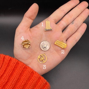 May include: Four gold-toned metal charms labeled 1 through 4. Charm 1 is a United Nations building, 2 is a rectangular bar, 3 is a circular lobster, and 4 is a typewriter. A US dime is included for scale, showing the charms' relative size.