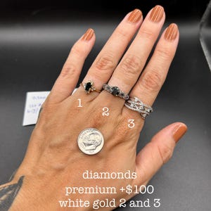 May include: Three rings worn on fingers. Ring 1 is a gold band with a black oval stone and small clear stones. Ring 2 is a silver-toned band with a black oval stone and filigree details. Ring 3 is a silver-toned crossover band with clear stones.