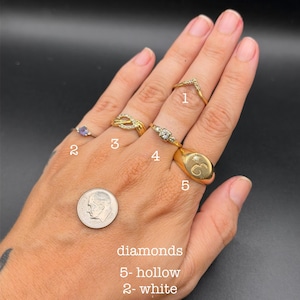 May include: Five gold rings numbered 1 through 5 are worn on fingers. Ring 1 is a chevron band, 2 is a white stone, 3 is a twisted band, 4 is a three-stone setting, and 5 is a signet ring with a small diamond. A US dime is visible for scale.